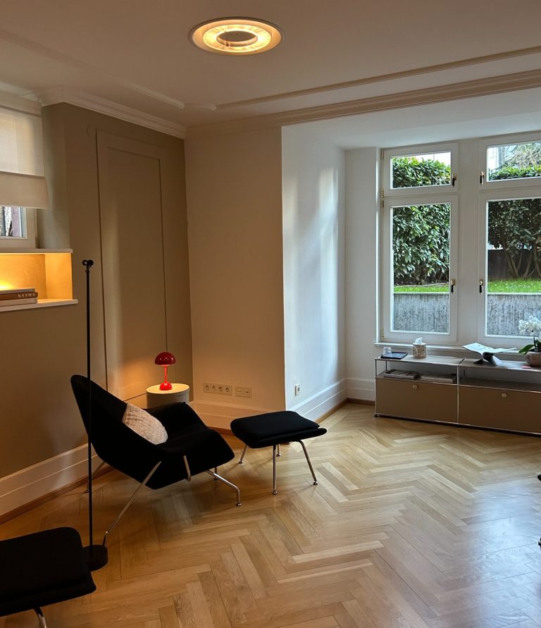 Heller Praxisraum mit Holzfußboden, Sessel, Tisch und Fenstern zur Gartenansicht.